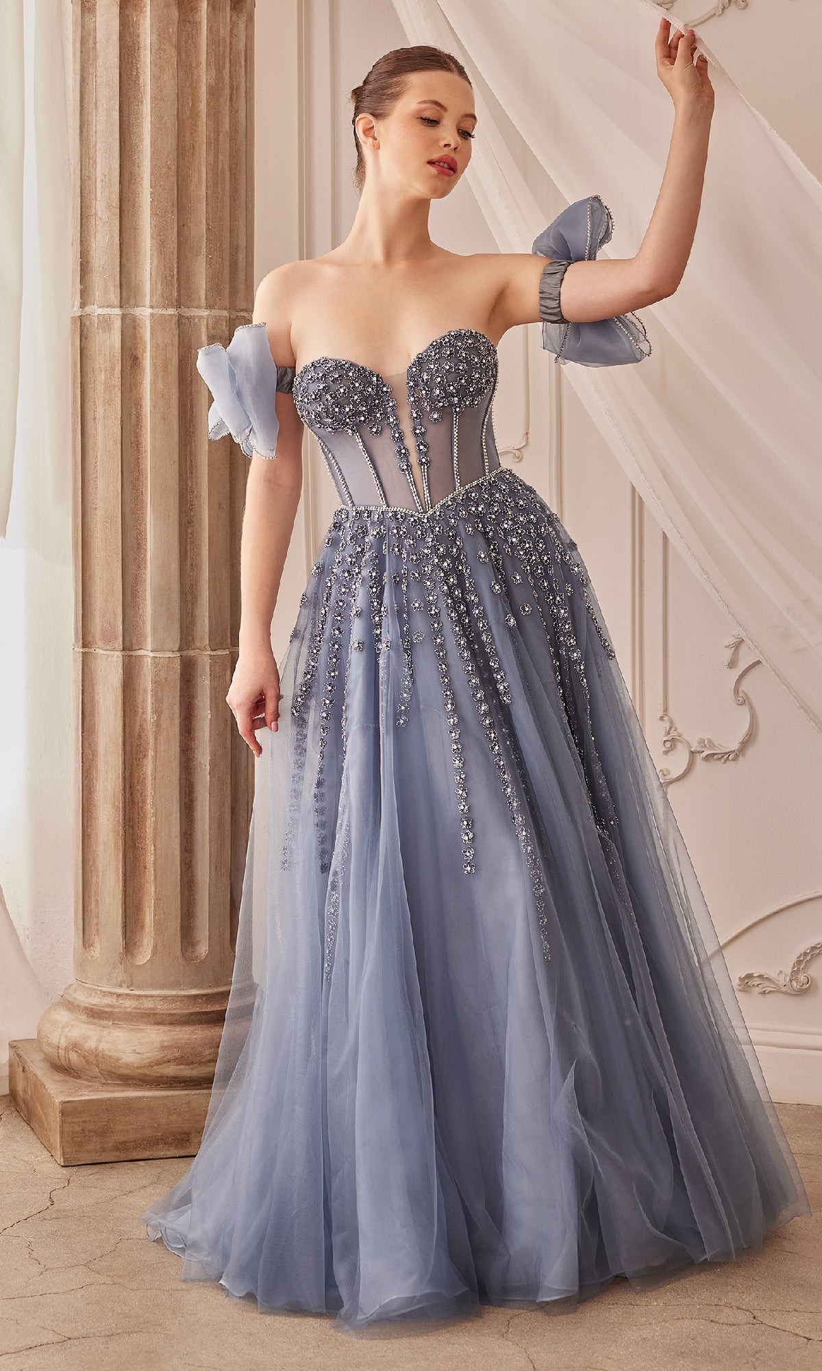 A woman stands indoors wearing the Strapless Long Prom Dress: Andrea and Leo A1338, a light blue beaded evening gown with tulle, posing near a column and draped curtains.